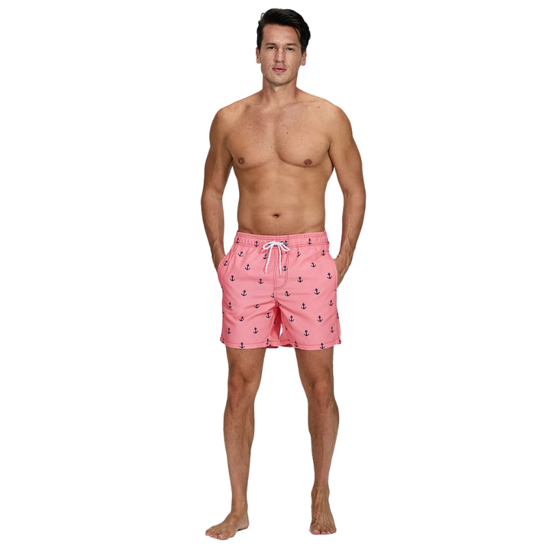 Man with shirt off and hands in pockets wearing pink swim shorts with a blue anchor pattern on white background.