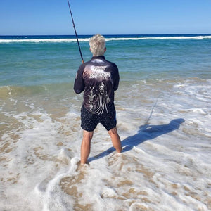 Man fishing on the beach wearing fishing shirt and beach shorts.